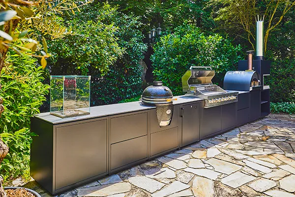 Foto der CB Outdoor Kitchen mit acht Modulen (Feuer-Modul, zwei Schubladen-Module, Monolith-Modul, Kühlschrank-Modul, Gasgrill-Modul, Pizzaofen-Modul und Feuerstellen-Modul) auf einer Terasse in einem Garten.